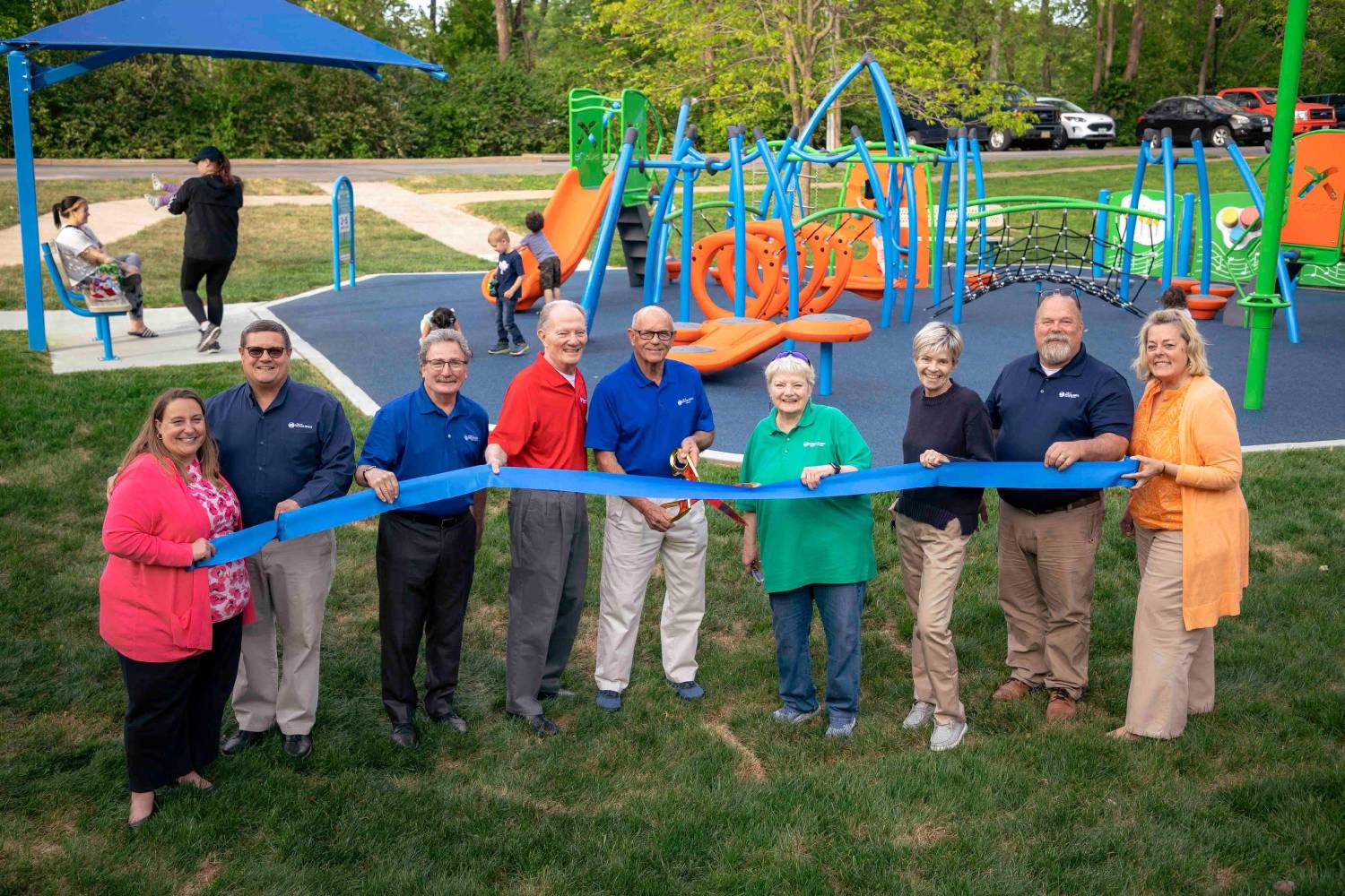The Mayor, City Administrator, Director of Parks and Recreation, and Members of the Maryland Heights City Council pose with a ribbon and oversized pair of scissors at an playground located in Parkwood Park. Behind them, various children play on the playground equipment.